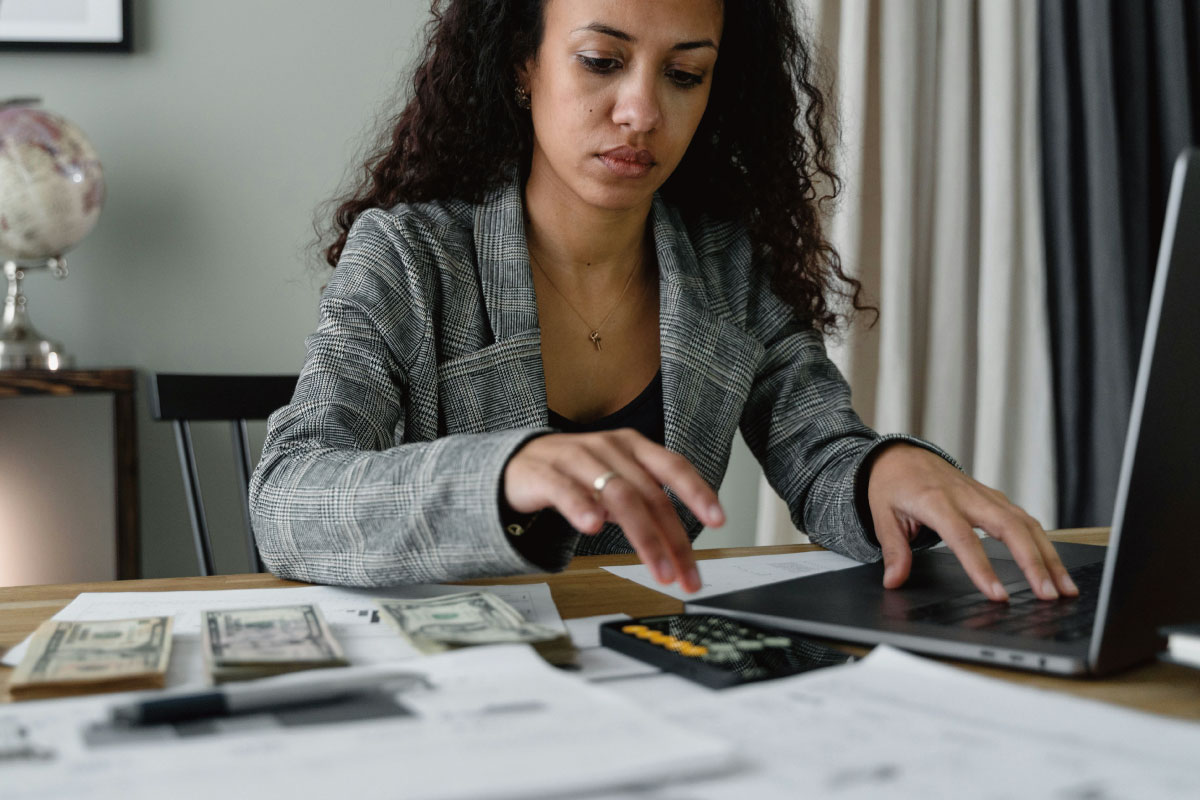 A woman sitting at a table with a computer, a calculator, and US dollars, seemingly calculating bills